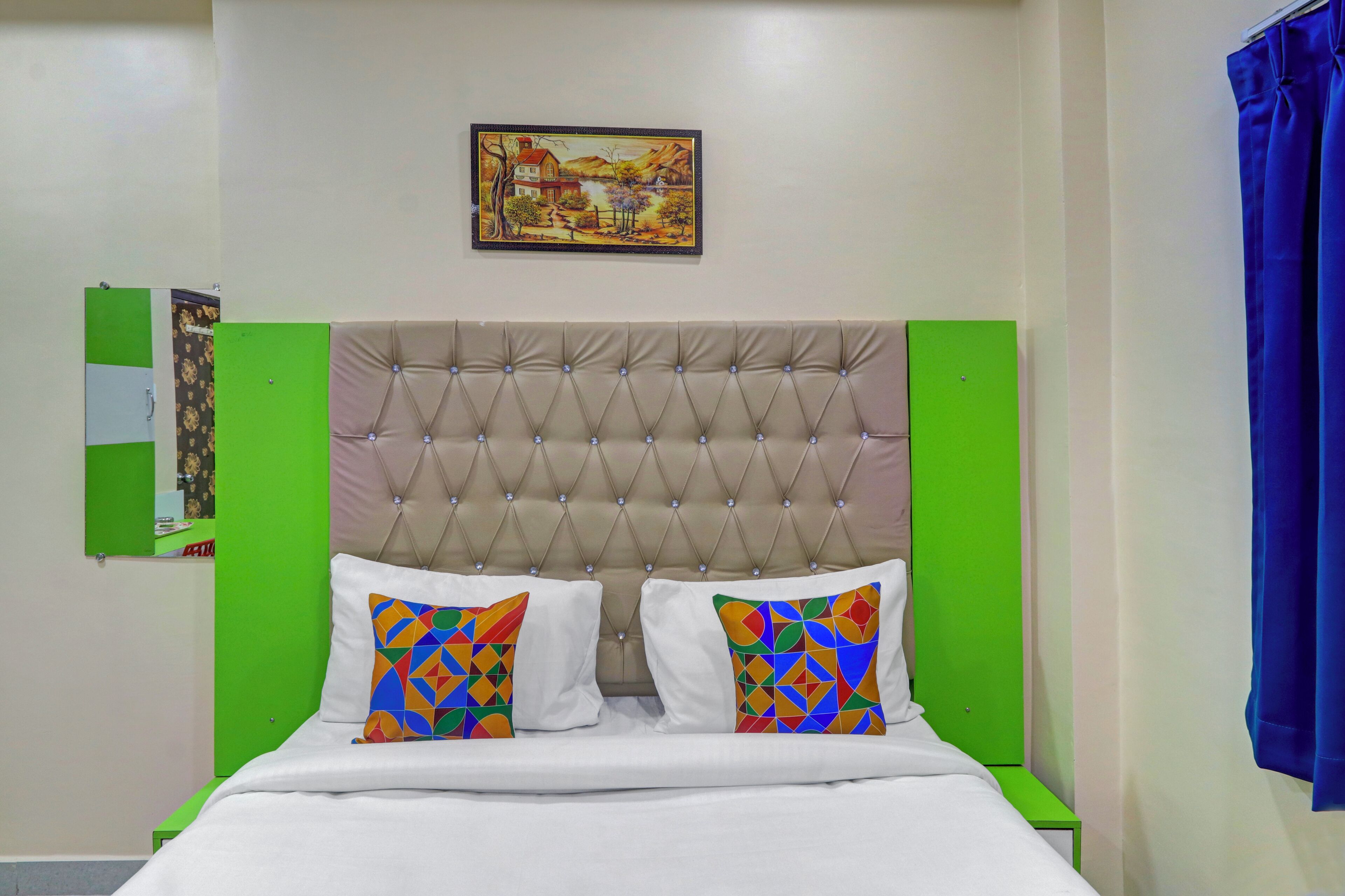 A hotel room with a tufted headboard, two colorful pillows, a framed picture on the wall, and a blue curtain.