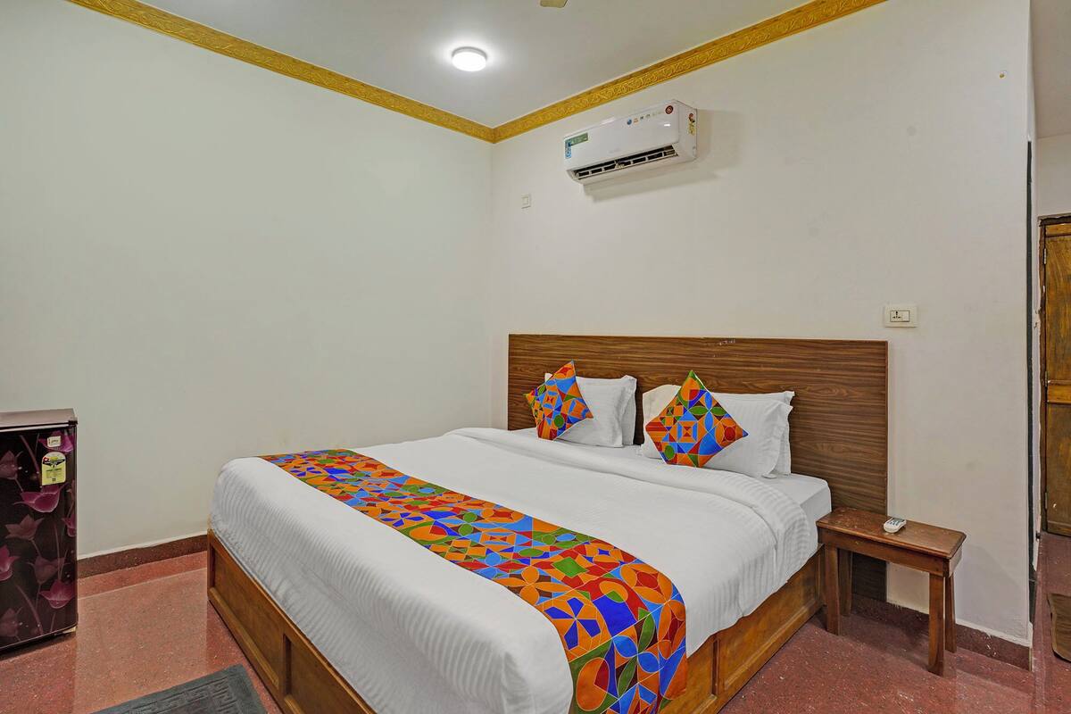 A bedroom with a wooden headboard, a bed with white linens, a colorful geometric-patterned bedspread, two decorative pillows, a bedside table, and an air conditioning unit on the wall.