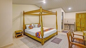 Un lit à baldaquin surmonté d’une couvre-lit aux motifs géométriques colorés, une petite table de chevet en bois et une chaise dans une pièce dont le sol est recouvert de carreaux décoratifs et dont le plafond est blanc.