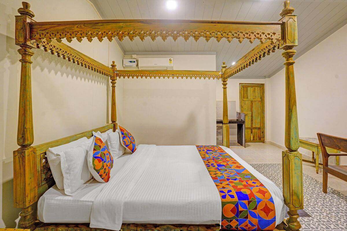 A four-poster canopy bed with white linens and colorful pillows, a wooden chair, and a patterned floor.
