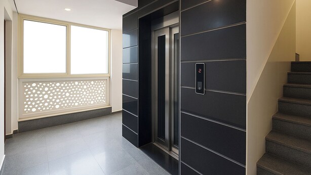 Un couloir moderne avec un escalier, une porte dotée d’un écran numérique et une fenêtre agrémentée d’un paravent décoratif.