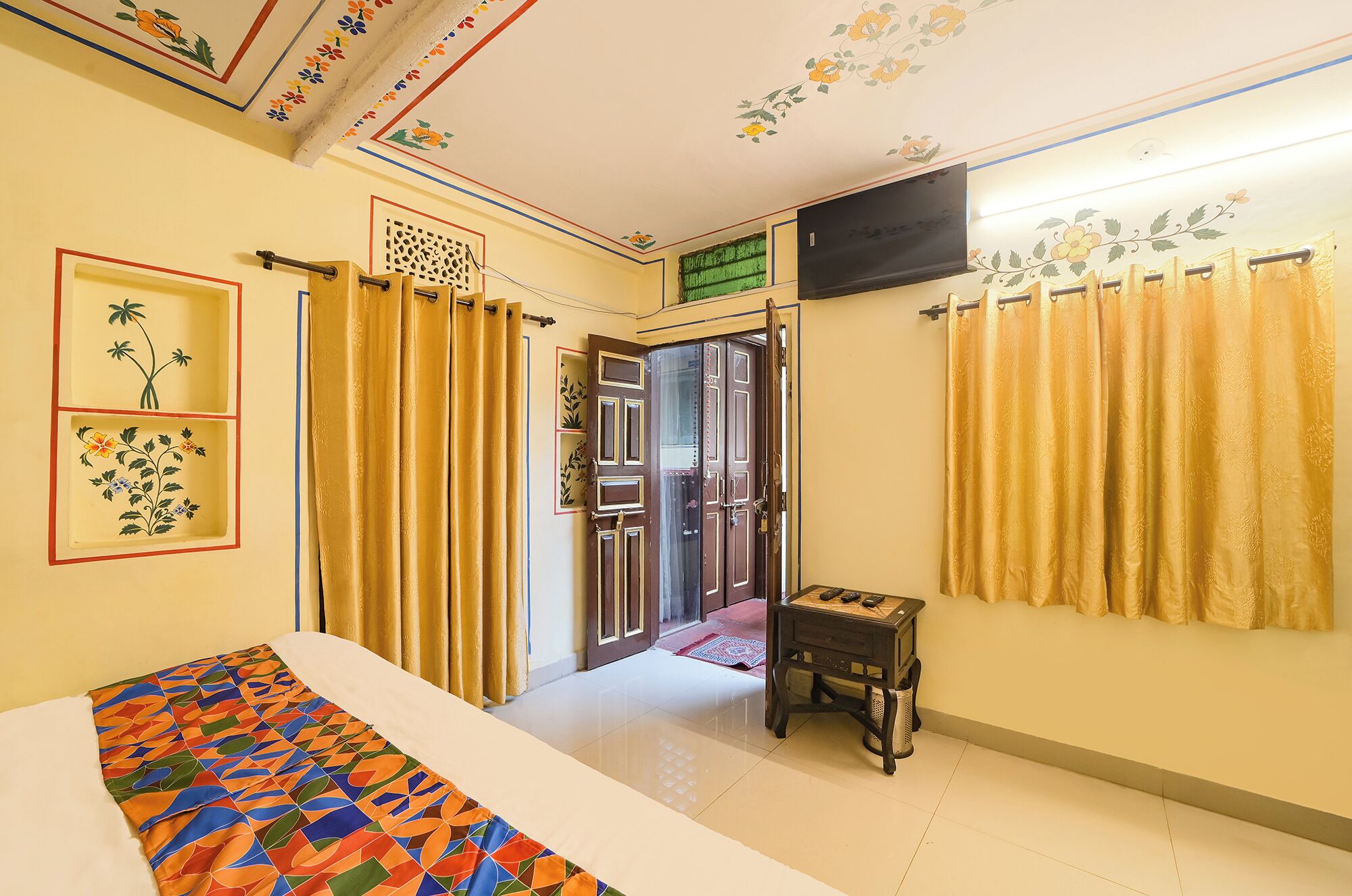 A room with a bed, a small table, a TV, and a door leading to another room.