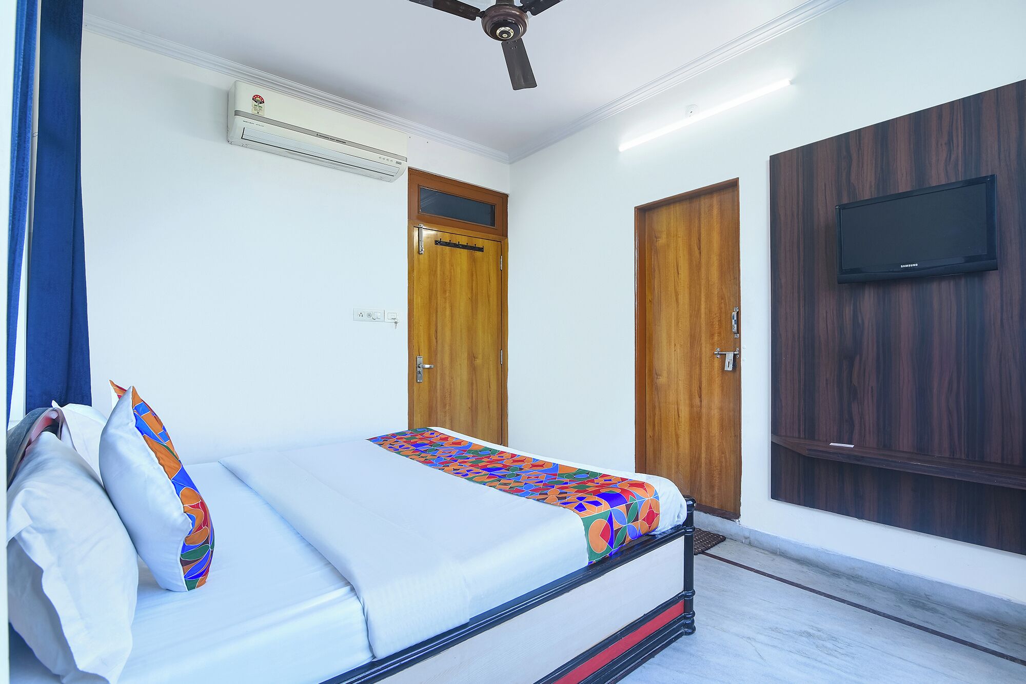 A bedroom with a bed, a ceiling fan, an air conditioning unit, two wooden doors, and a television mounted on the wall.