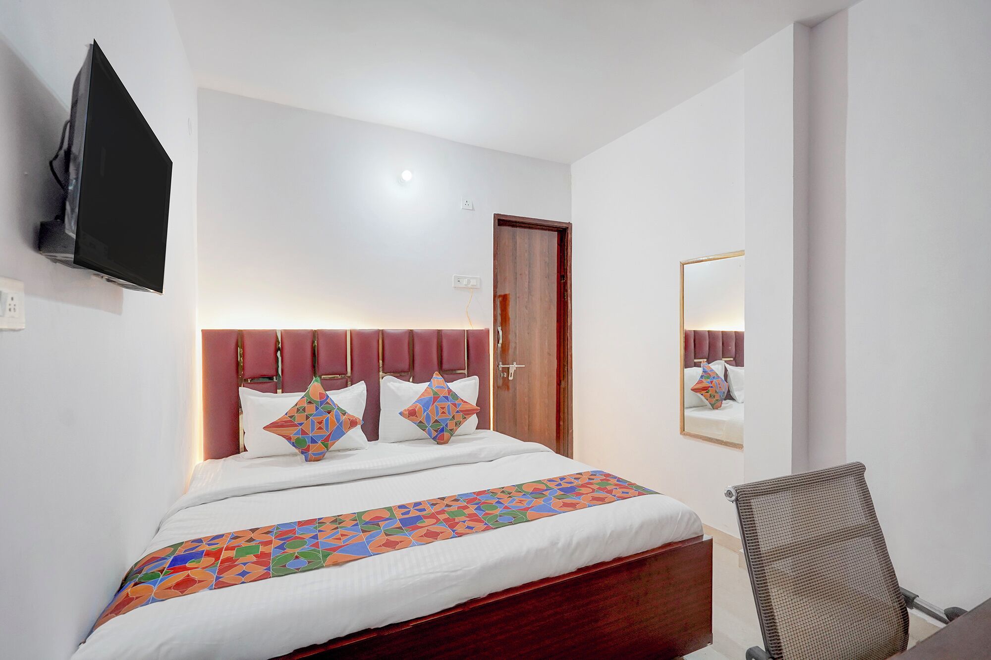 Una camera d'hotel con un letto, una televisione a parete, una porta, uno specchio e una sedia.