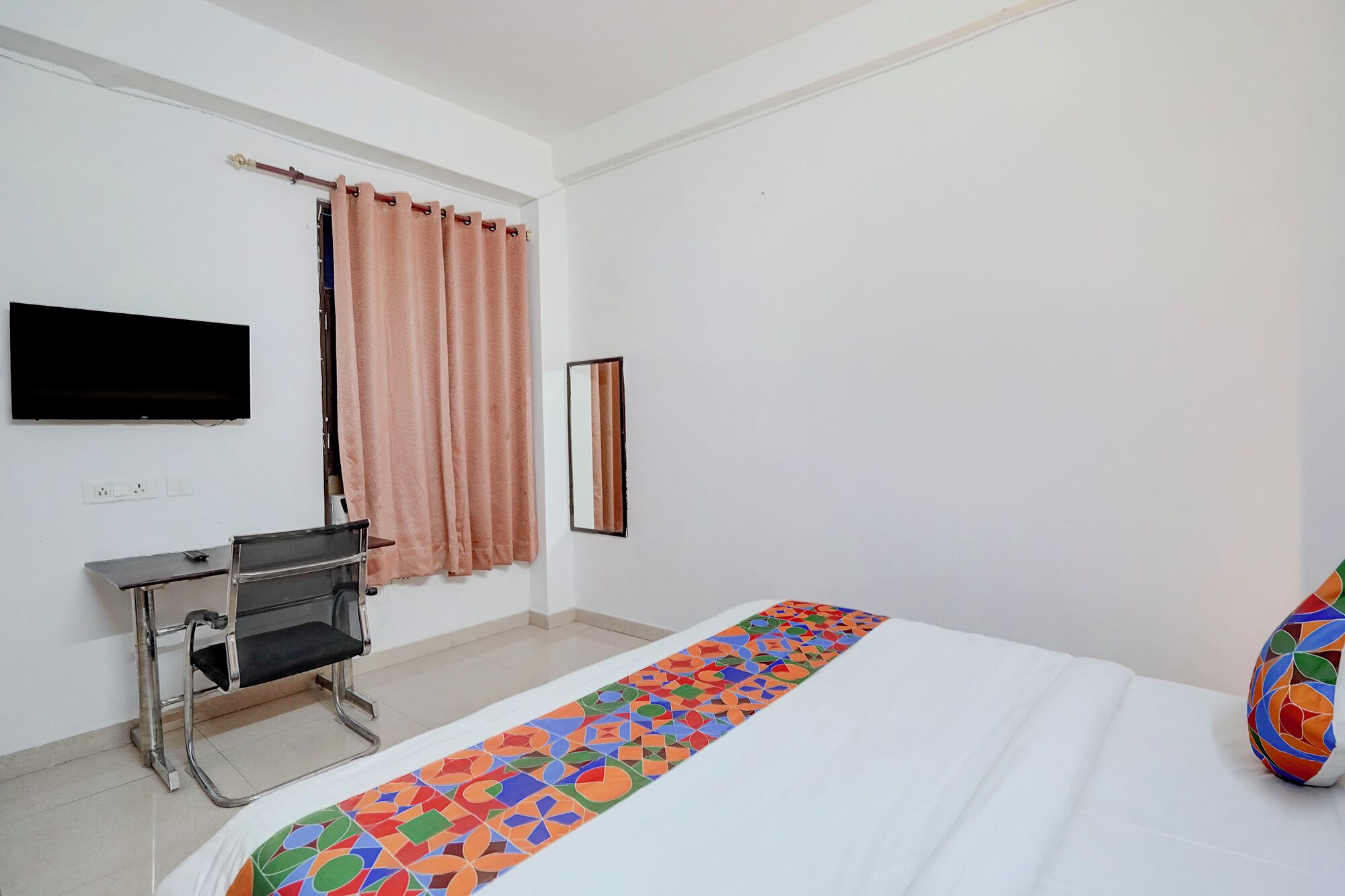 A hotel room with a bed, a desk with a chair, a television mounted on the wall, and a window with curtains.