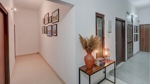 A hallway with a tiled floor, a wall-mounted picture frame, a small table with a vase and books, and a door with a glass panel. - Fabhotel Tree View Residency (Lucknow)