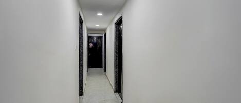 Um corredor estreito, bem iluminado, com paredes brancas e chão de azulejos, que leva a um aposento escuro no final.