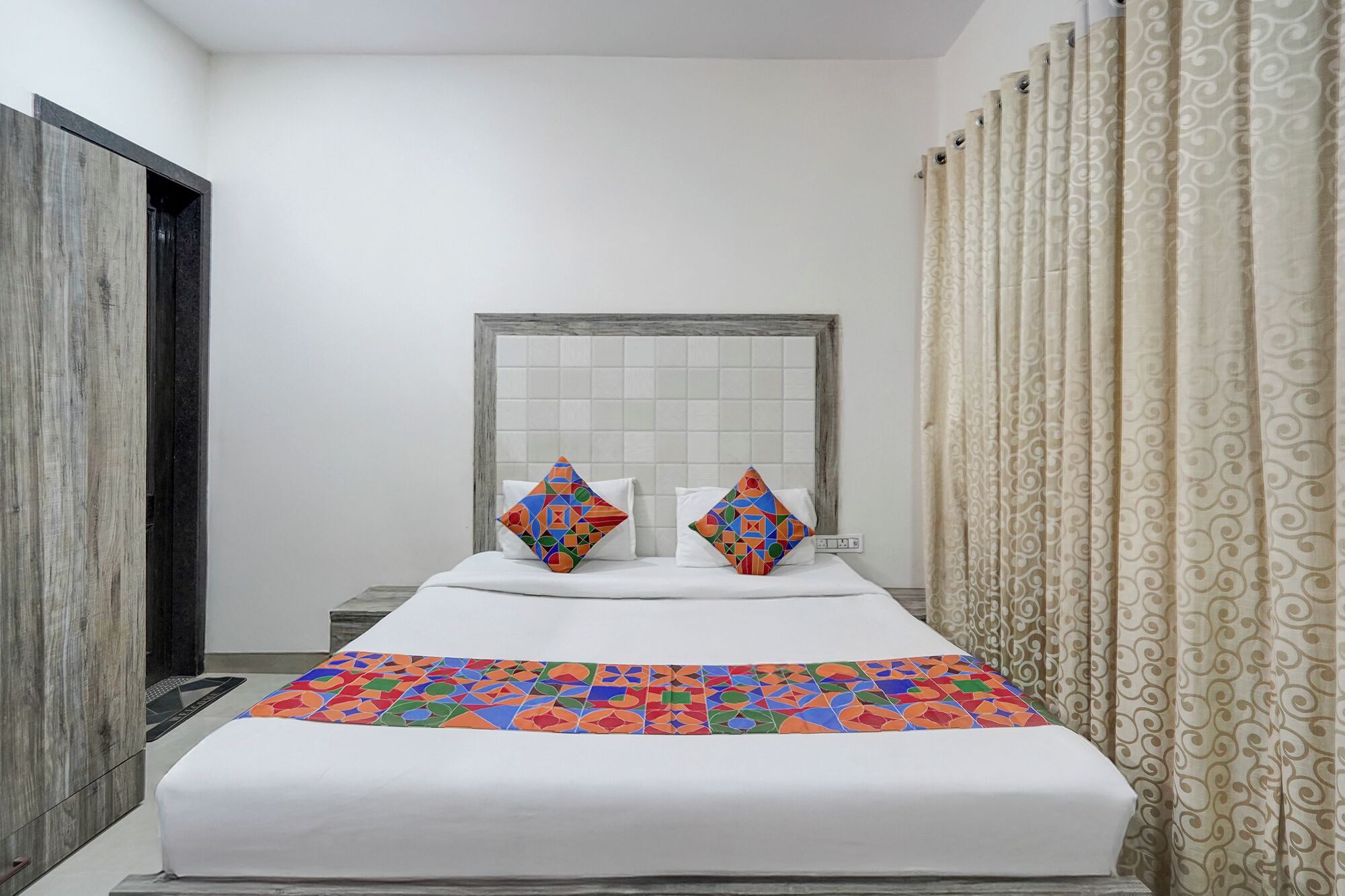 A hotel room with a bed, two pillows, a headboard, and a patterned curtain.