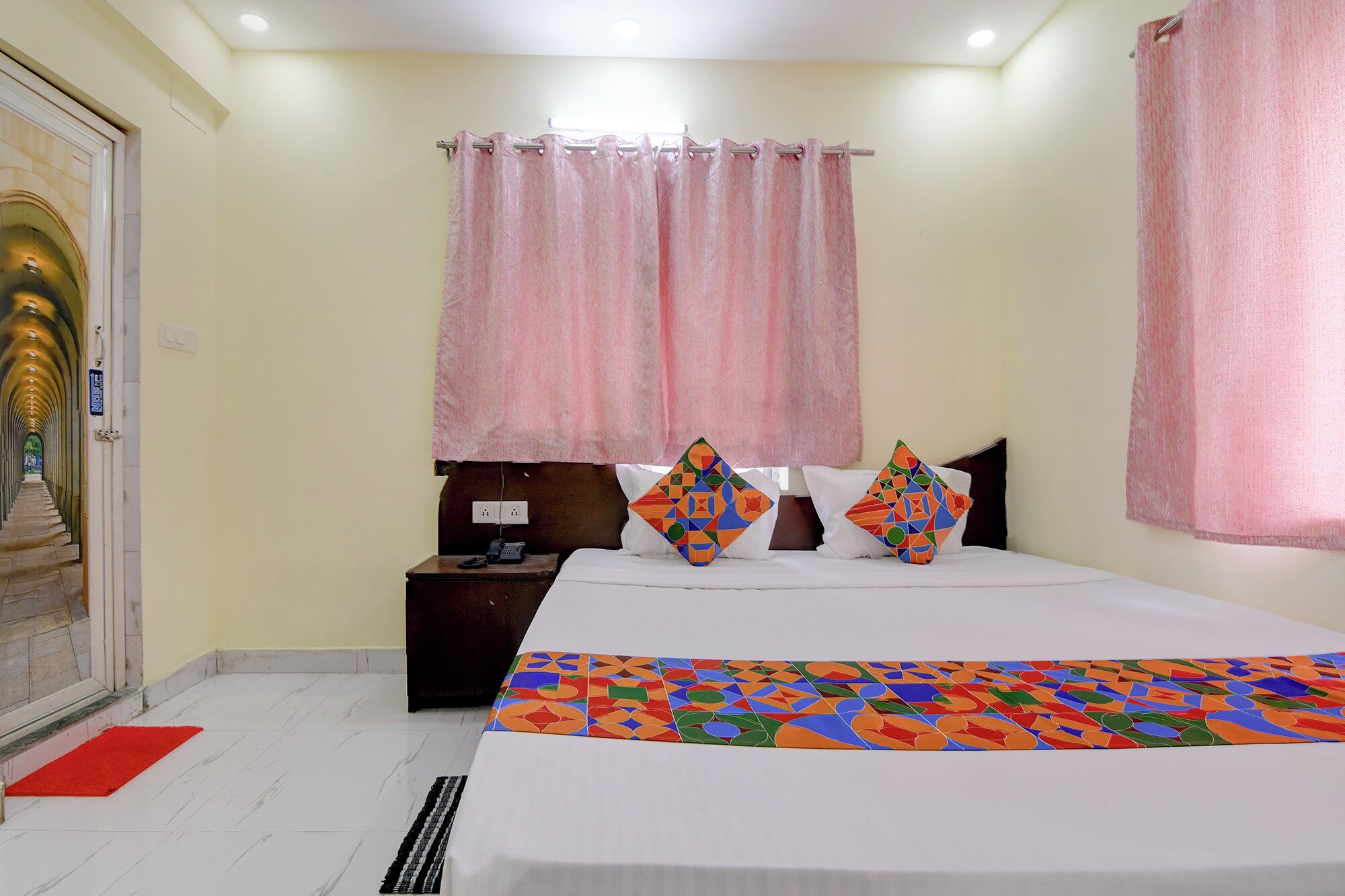 A hotel room with a bed, a nightstand, a red mat, and a view of a corridor with a door.