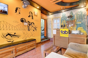 Een hotel lobby met een receptiebalie, een zithoek met een bank en een houten zitbank, en een muur met kunstzinnige muurschilderingen.