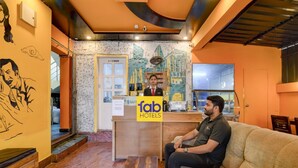 Un espace d’accueil d’hôtel comprenant un comptoir de réception, un canapé, une table basse en bois et un mur orné d’une fresque.