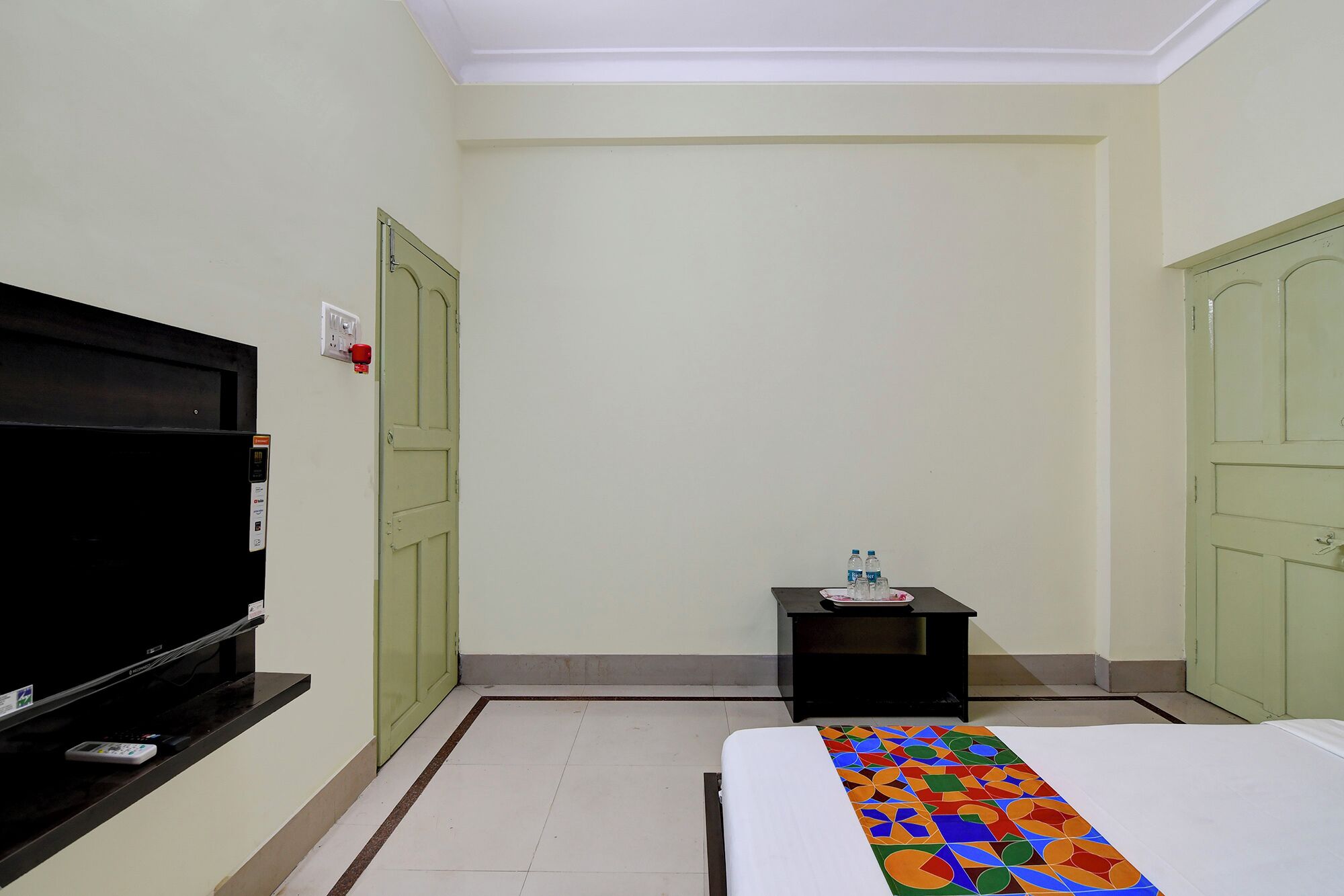 A hotel room with a bed, a television, a small table with water bottles, and two green doors.
