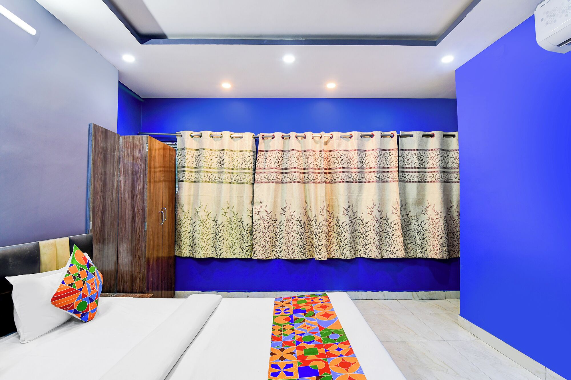 A bedroom with a bed, a colorful pillow, a blue wall, and a curtain with a floral pattern.