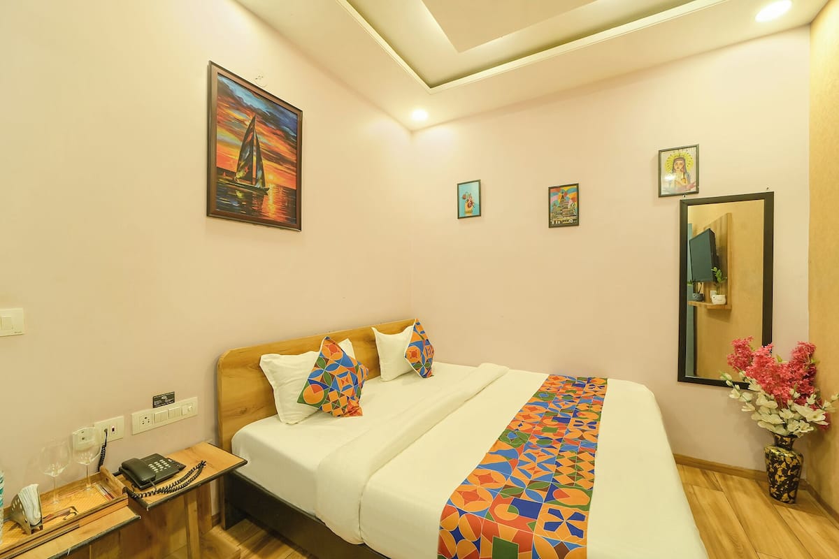 A hotel room with a bed, a bedside table with a phone and glasses, a mirror, and a wall with framed pictures.