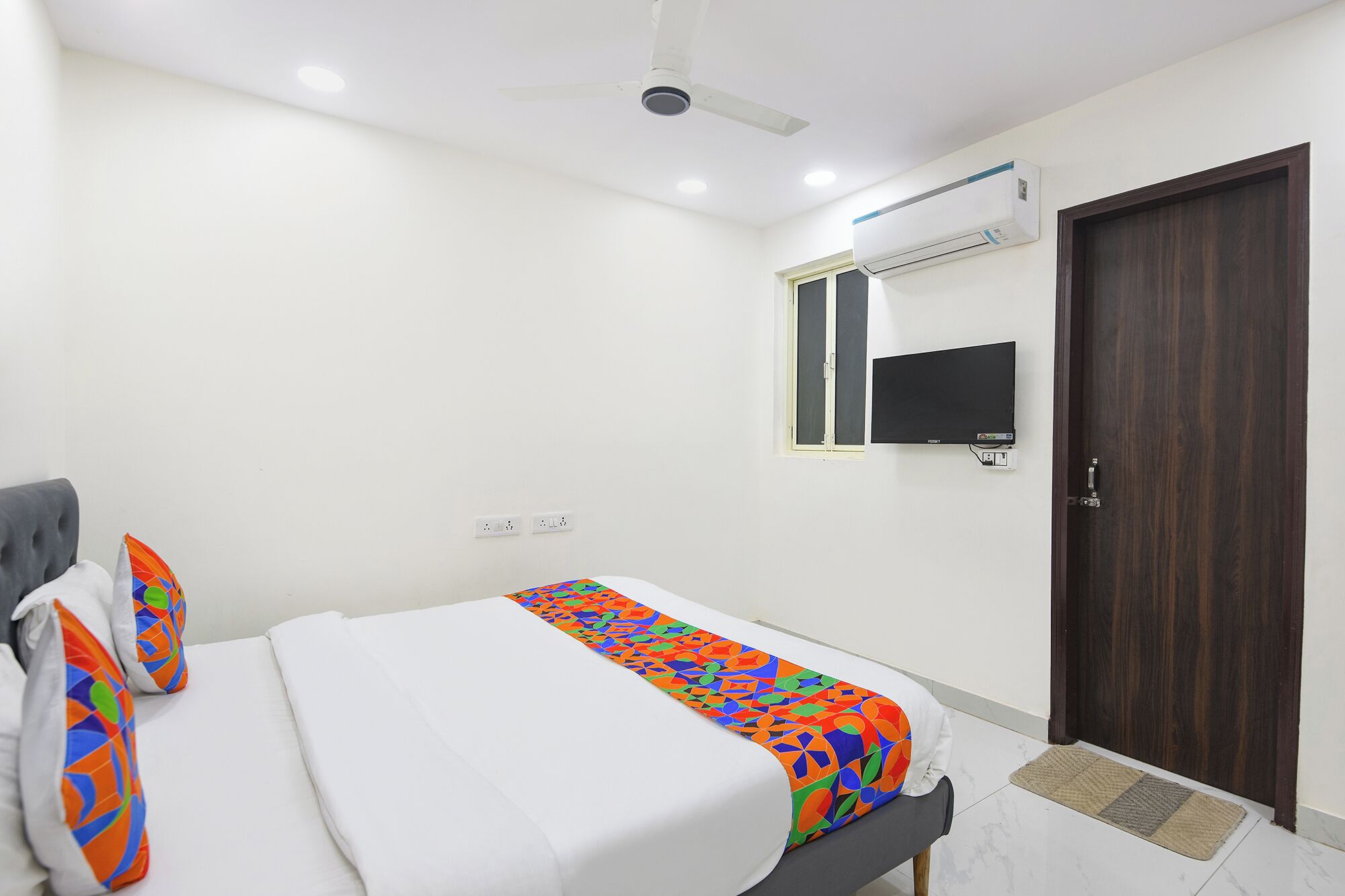 A bedroom with a bed, a ceiling fan, an air conditioning unit, a TV mounted on the wall, and a wooden door.