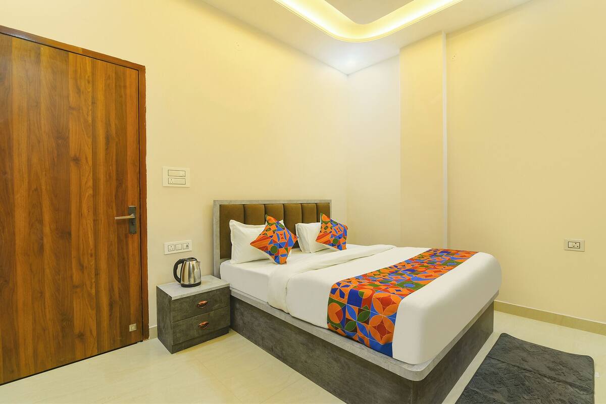 A bedroom with a wooden door, a bed with a patterned blanket, a bedside table with a kettle, and a small rug.