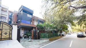 A gated entrance with a blue sign, a paved sidewalk, and a street with parked cars. - Via Esscon House (Gurugram)