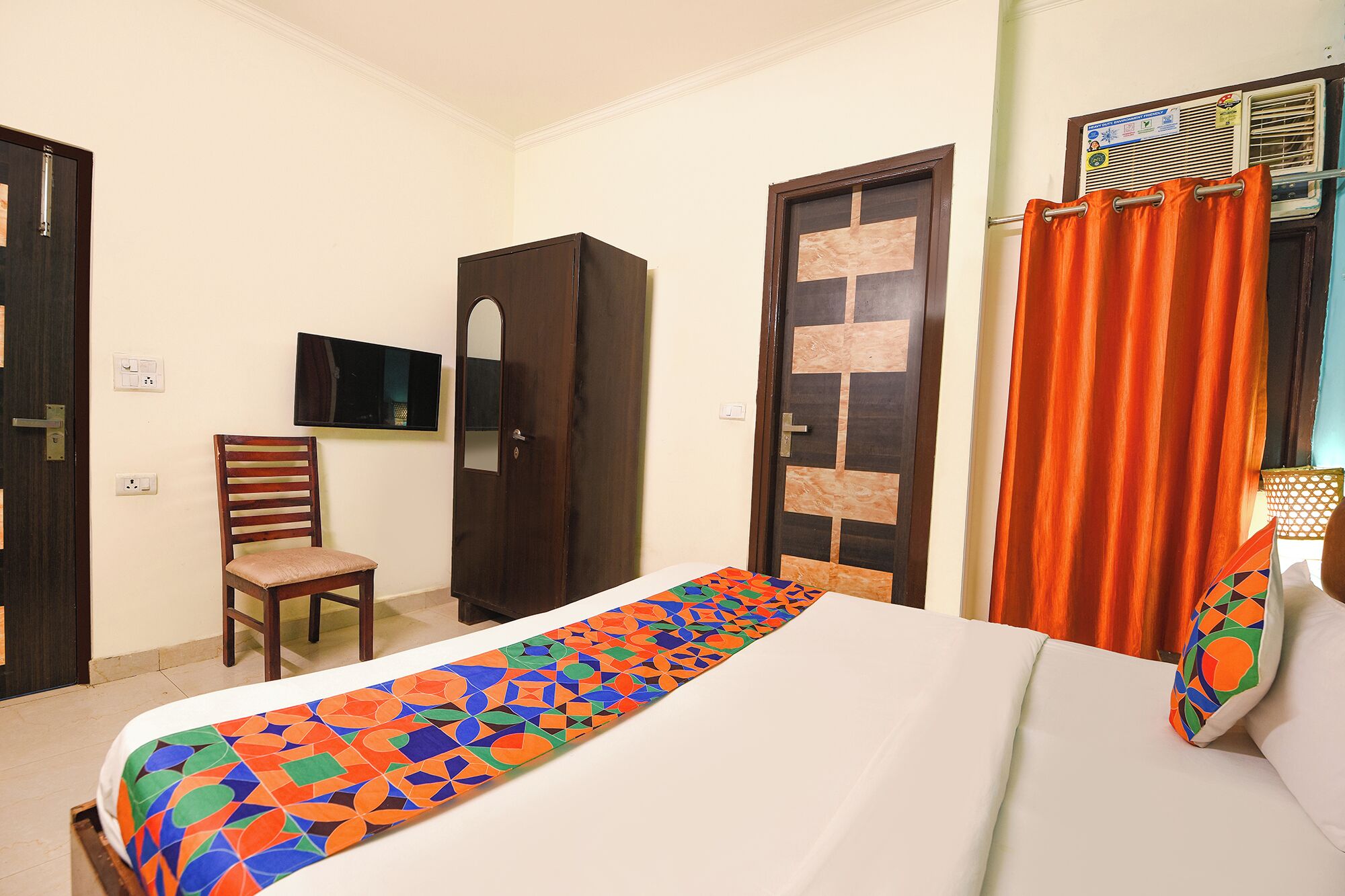 Una habitación de hotel con cama, una silla, armario, televisor y ventana con cortinas.