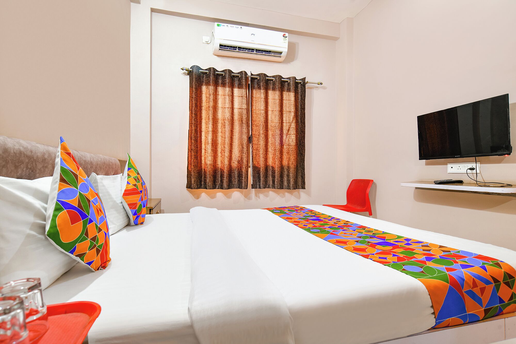 A hotel room with a bed, a television, an air conditioning unit, a window with curtains, and a red chair.