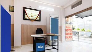 A reception desk with a 'Via' sign, a framed picture with a garland, a 'Welcome Hotel Kanha Palace' sign, and a view of the outside. - Via Kanha Palace (Ujjain)