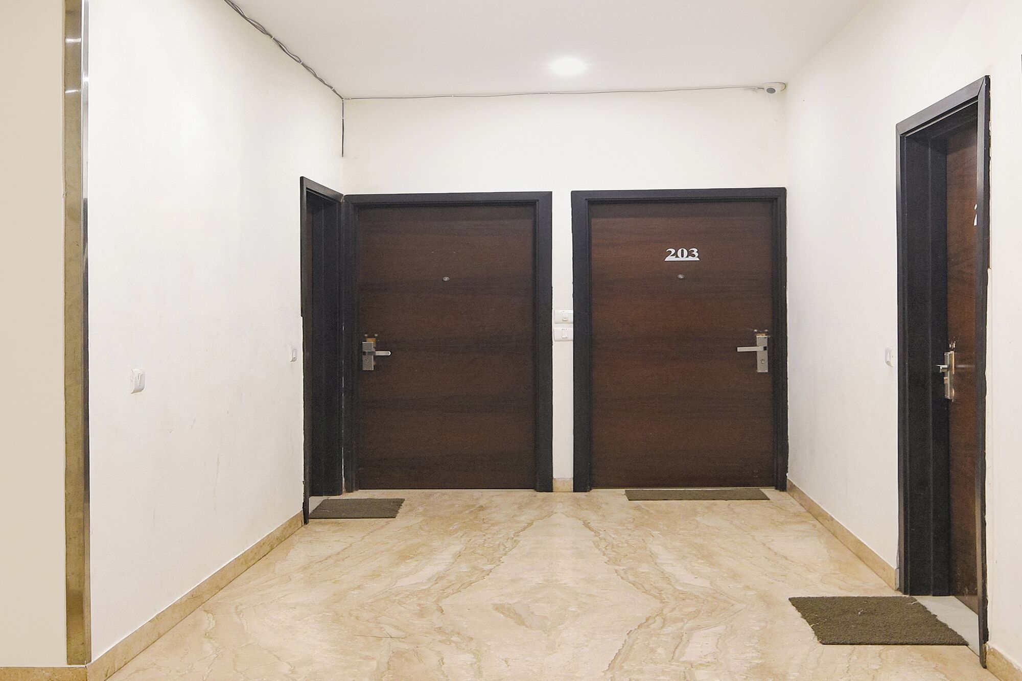 A hallway with two dark wooden doors, each marked with the number 203. The floor is tiled with beige marble, and there are small mats at the doors' entrances.