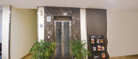 An elevator with a marble wall, two potted plants, and a rack with magazines.