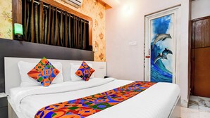 Quarto de hotel com cama, edredom colorido estampado com padrão geométrico, cabeceira branca, parede com papel de parede floral, porta com mural de golfinhos e janela com cortinas.