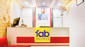 Uma área de recepção com um sofá vermelho, um balcão de recepção com a sinalização 'fab hotels', uma estátua de Buda e um ventilador fixado na parede.