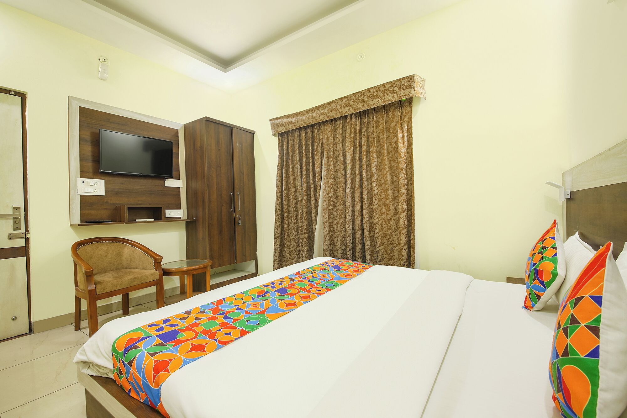 A hotel room with a bed, a television mounted on the wall, a wardrobe, a chair, and a window with curtains.