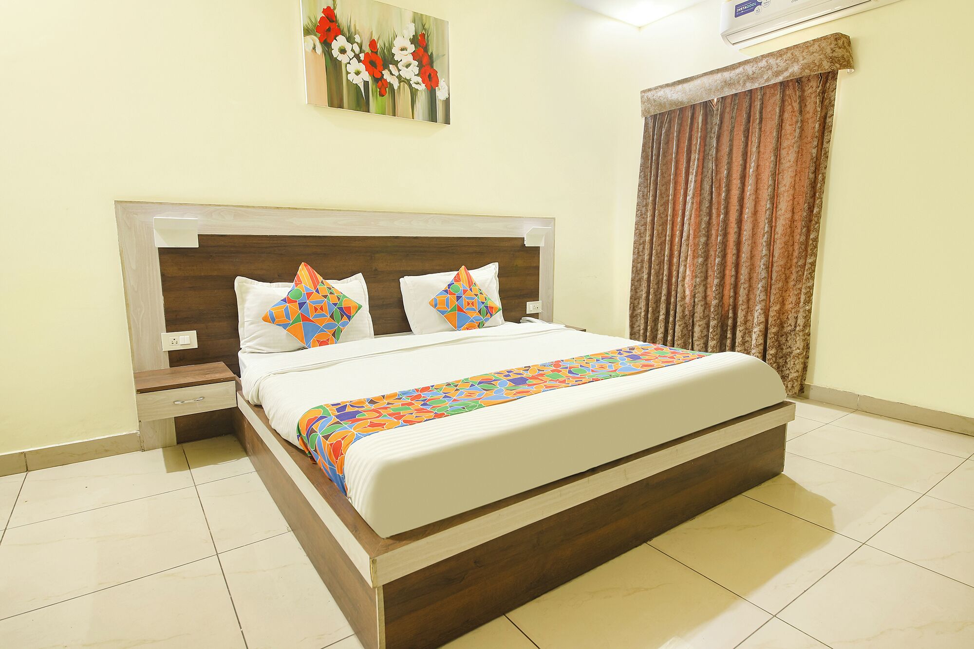 A hotel room with a bed, a nightstand, a wall-mounted air conditioner, and a window with curtains.