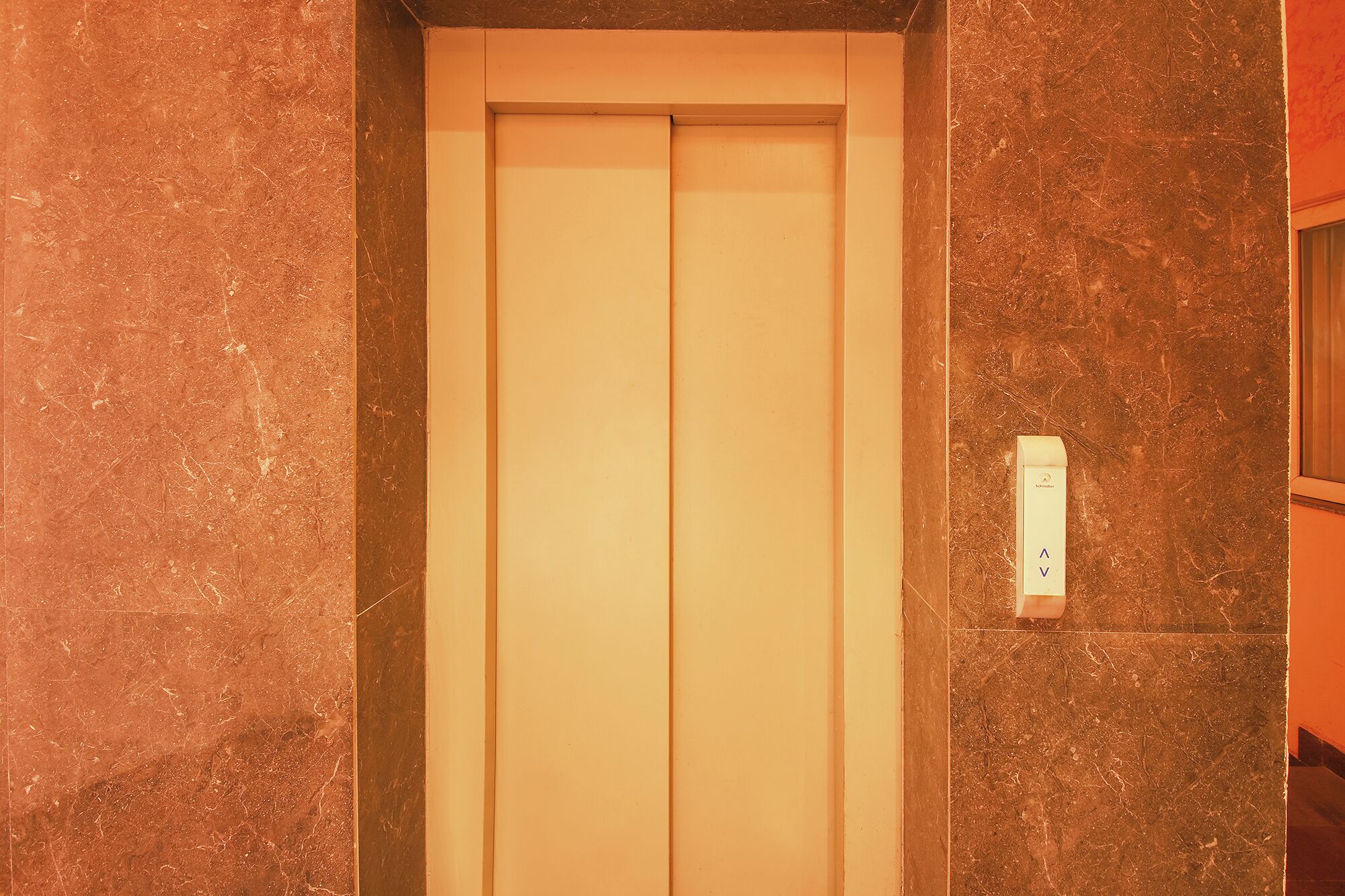 Een gesloten liftdeur met een marmeren muur en een lichtschakelaar.