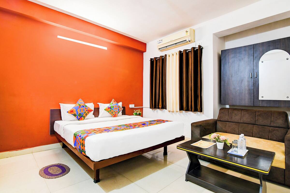 A hotel room with a bed, a sofa, a small table, and an air conditioning unit.