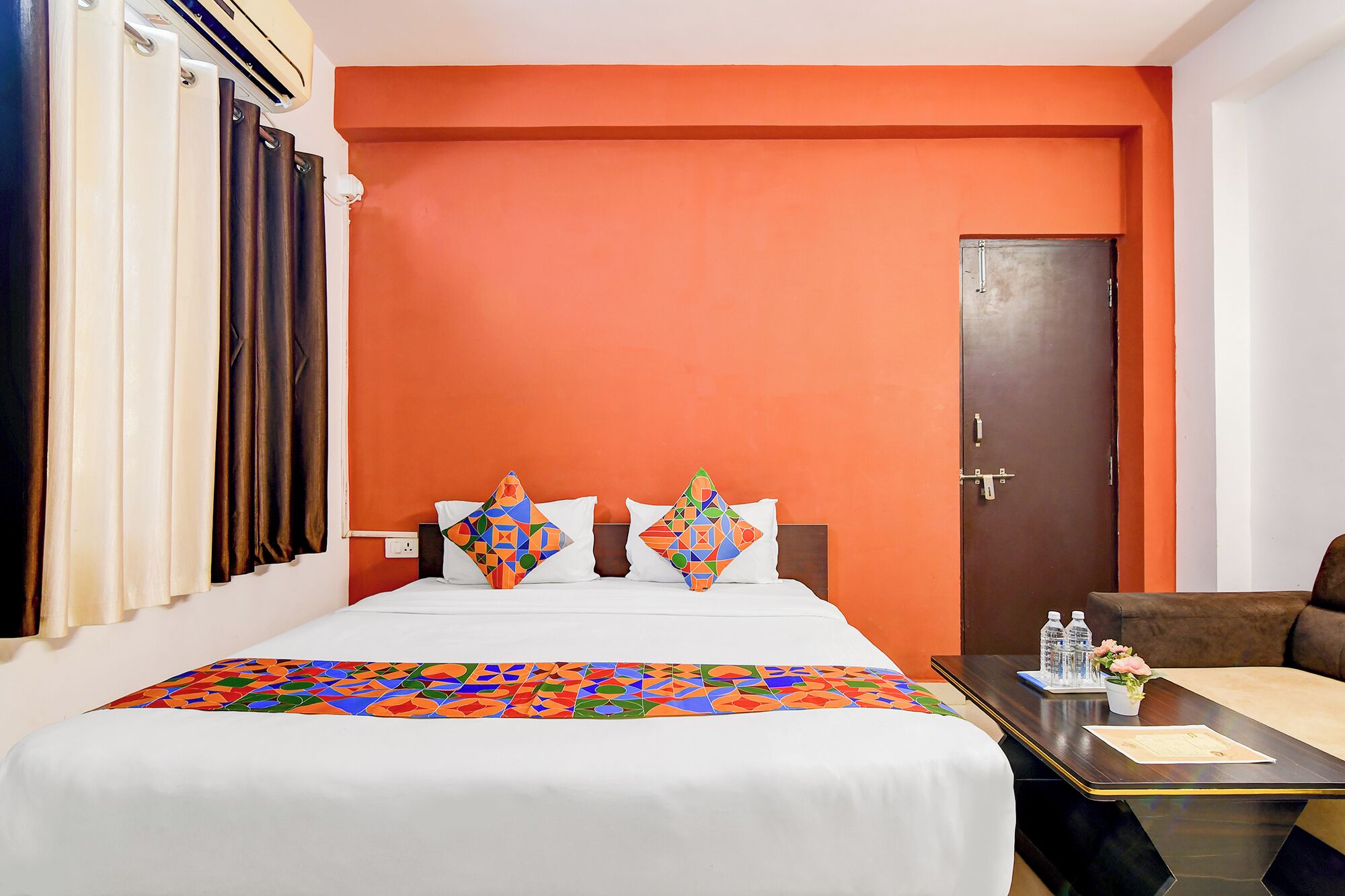 A hotel room with a bed, two pillows, a nightstand, a door, and a window with curtains.