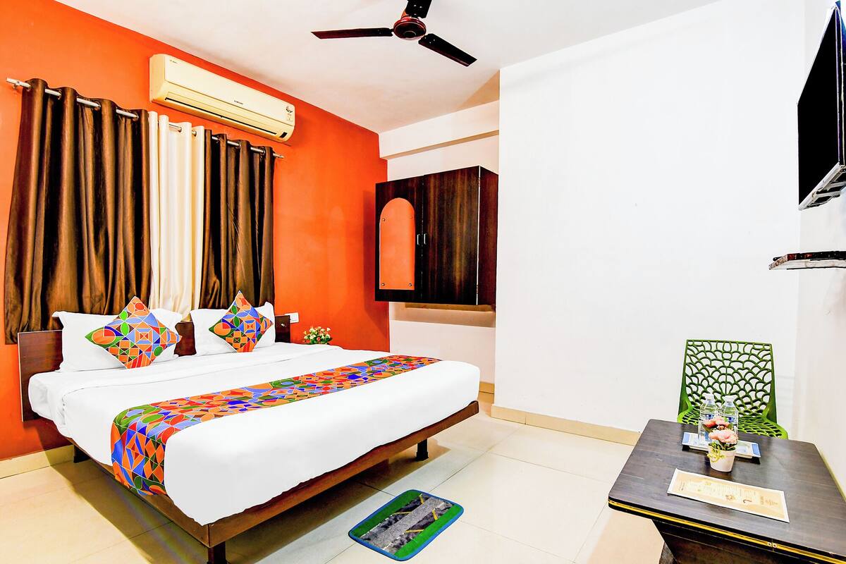 A hotel room with a bed, curtains, an air conditioning unit, a ceiling fan, a wooden cabinet, a small table with a vase, and a green chair.
