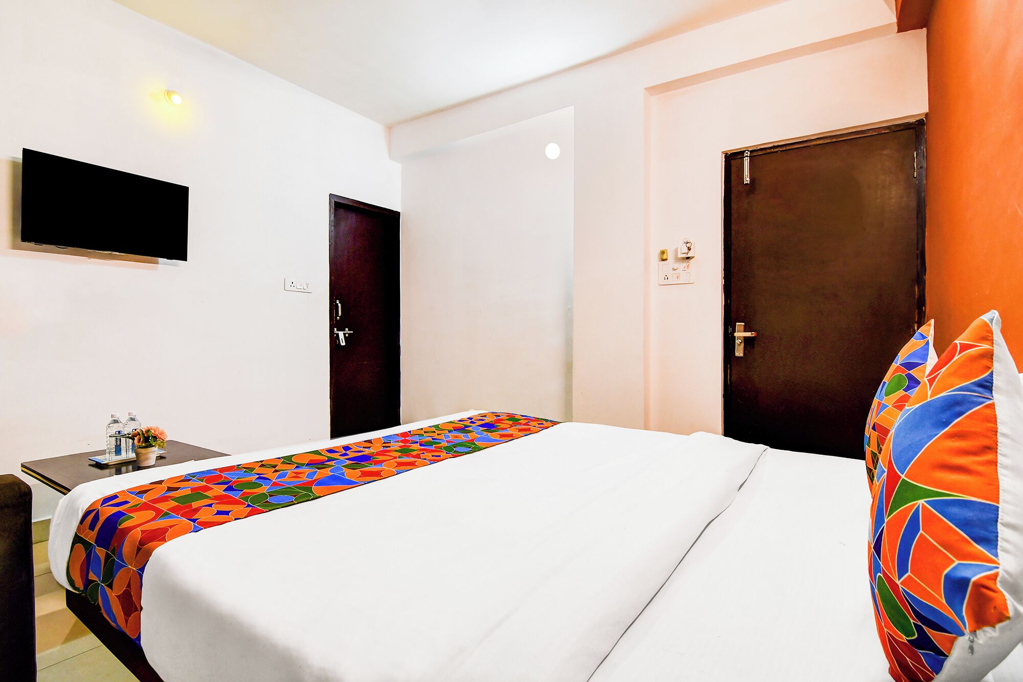 Habitación de hotel con una cama, un escritorio con silla, un televisor montado en la pared y dos puertas.
