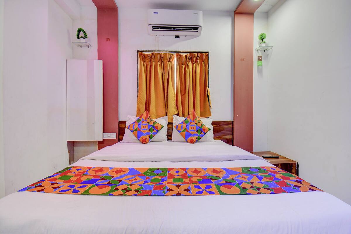A hotel room with a bed, a nightstand, an air conditioning unit, a window with curtains, and a wall-mounted shelf with a plant.