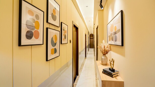 A hallway with framed abstract art on the walls, a wooden console table with books and a vase of dried flowers, and a door at the end. - Fabhotel Jk (Mumbai)