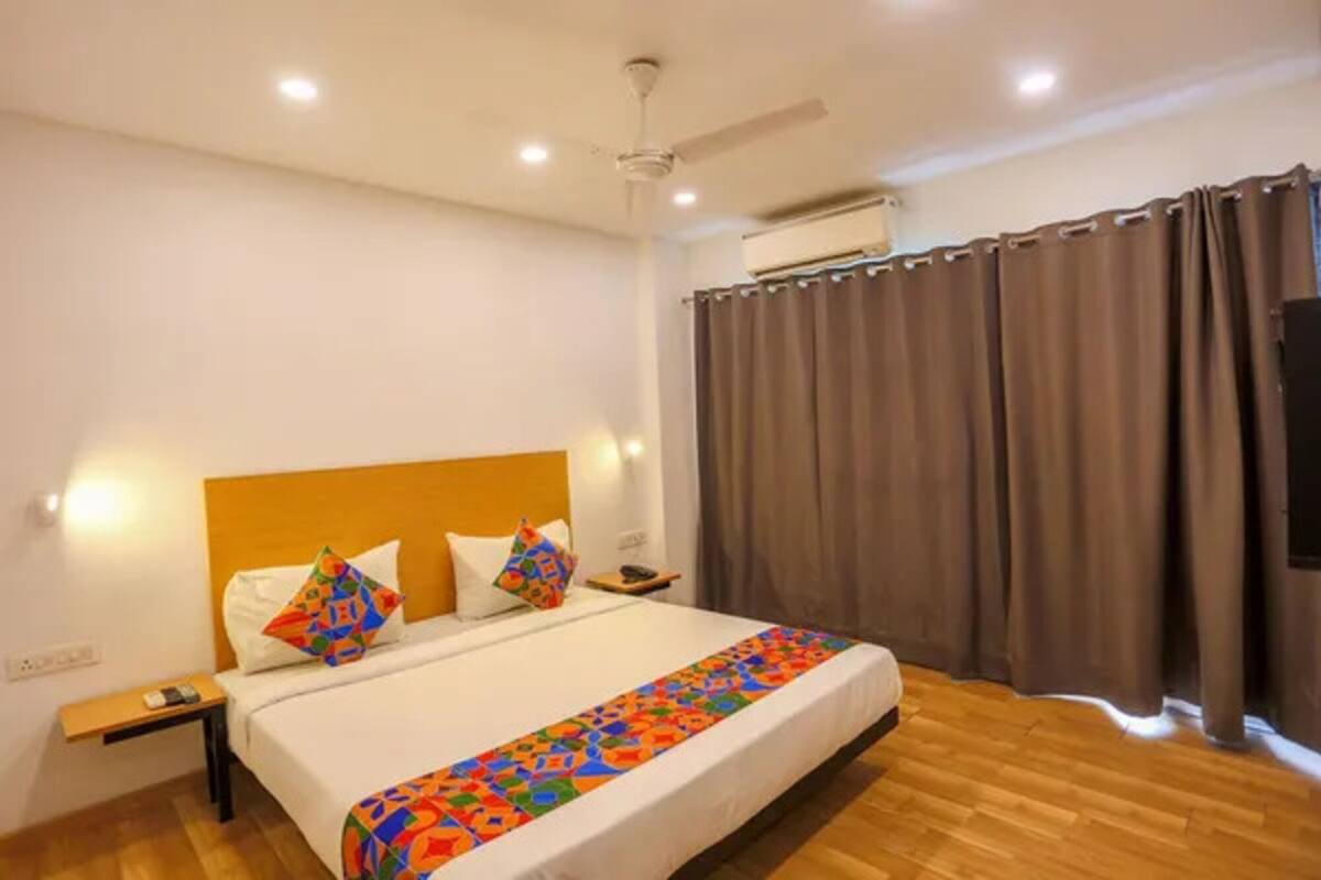 A hotel room with a wooden floor, a bed with a colorful blanket, a headboard, a bedside table with a phone, and a window with curtains.