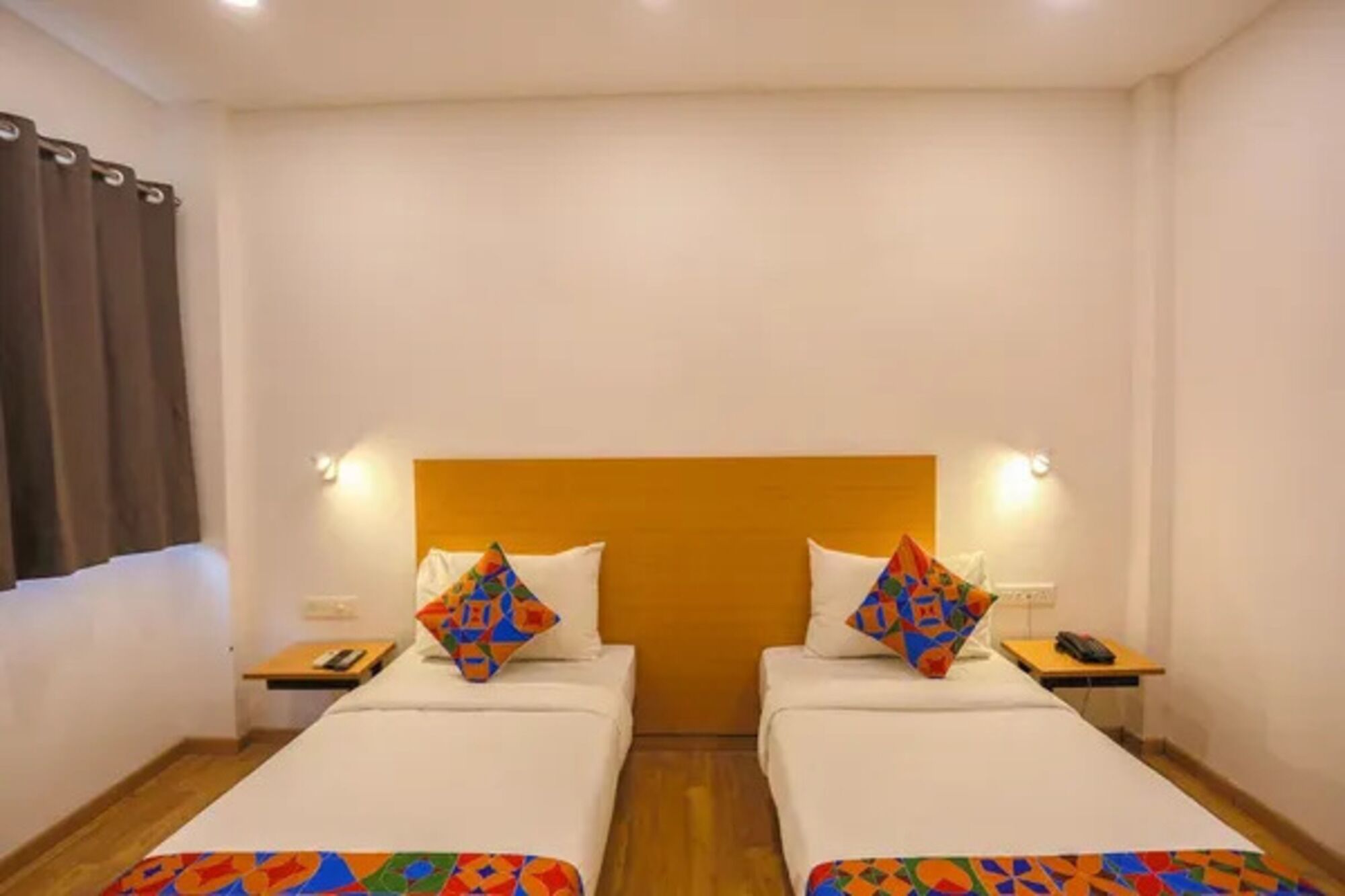 Une chambre d’hôtel avec deux lits simples, chacun agrémenté d’un coussin aux motifs géométriques et aux couleurs vives. De chaque côté des lits, on trouve une table de chevet avec une lampe et un téléphone.