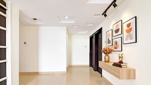 A hallway with a wooden console table displaying books and decorative items, a white wall with framed artwork, and recessed lighting on the ceiling. - Fabhotel Park Street Inn (Ranchi)