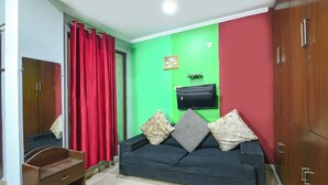 A room with a green wall, a red curtain, a black sofa with patterned cushions, a wooden cabinet, and a flat-screen TV mounted on the wall. - Via Elite Stay (Gurugram)