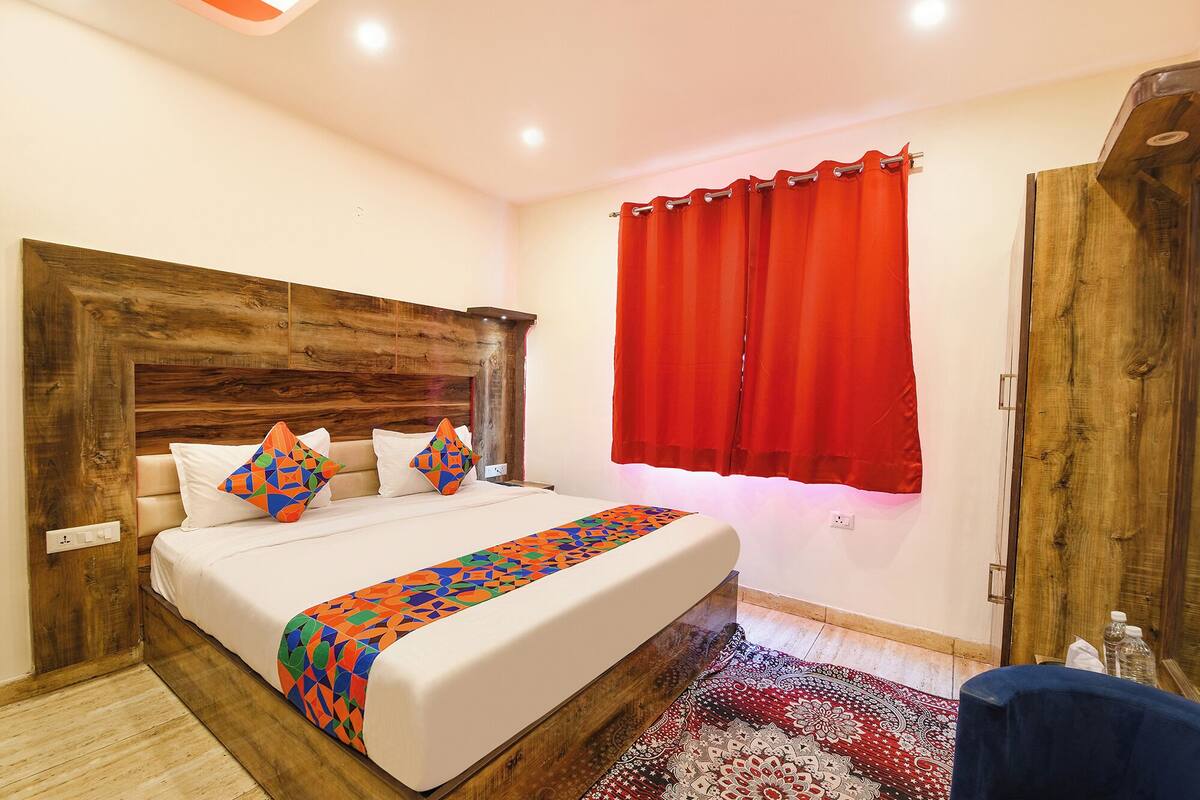 A bedroom with a wooden headboard, a bed with white bedding and colorful geometric pillows, a red curtain, and a patterned rug.