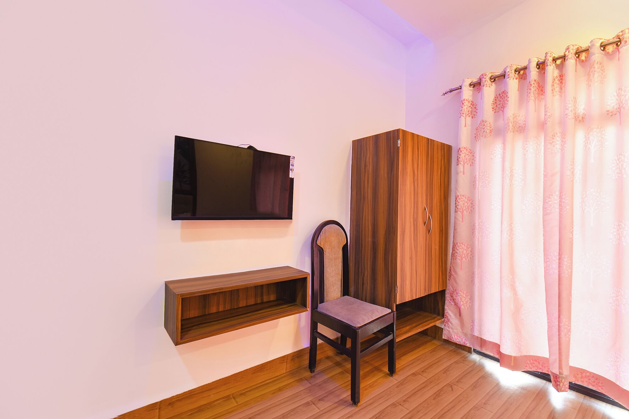 A room with a TV mounted on the wall, a wooden wardrobe, a chair, a small wooden shelf, and a window with pink curtains.