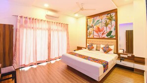 Habitación de hotel con cama, ventilador de techo, aire acondicionado, suelo de madera, ventana con cortinas y un cuadro floral en la pared.