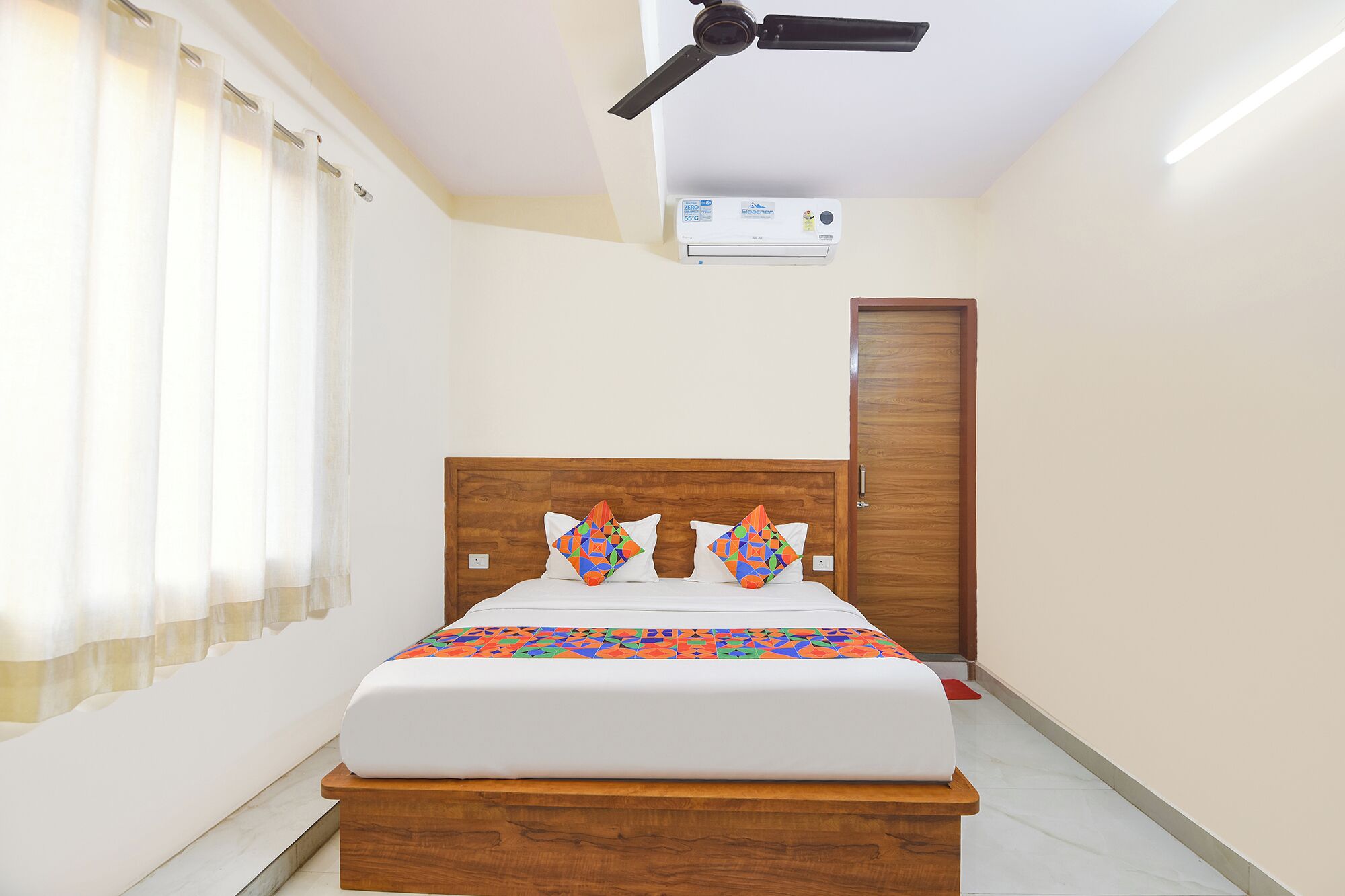 A hotel room with a wooden headboard, a bed with white bedding and colorful geometric-patterned pillows, a ceiling fan, an air conditioning unit, and a wooden door.