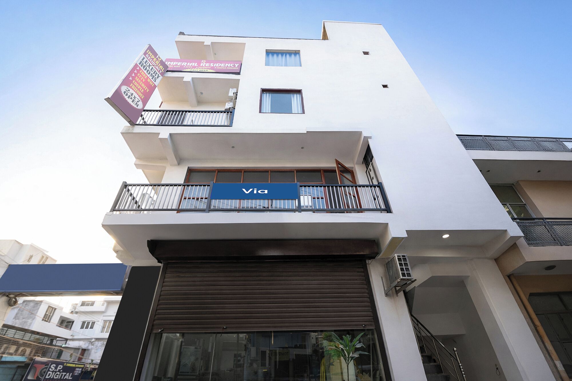 A multi-story building with a blue sign reading 'VIA' on the ground floor, a balcony with a metal railing, and a closed shutter on the ground floor.