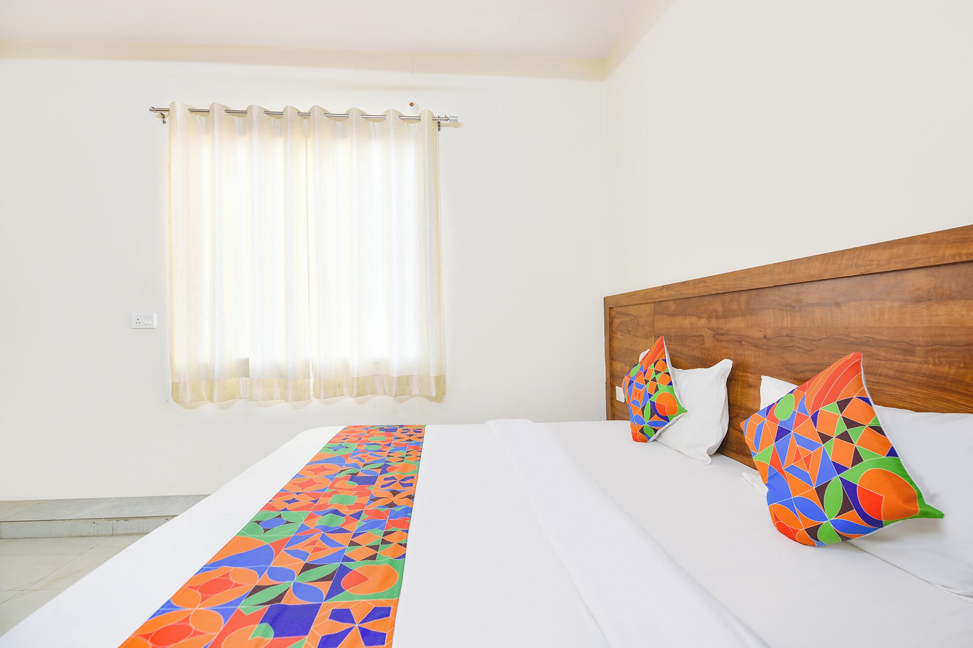 A hotel room with a bed, a wooden headboard, a window with curtains, and colorful pillows.