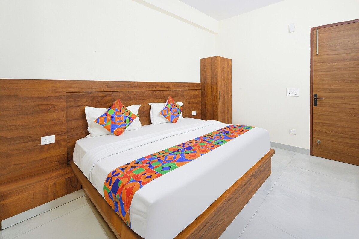 A neatly made bed with a colorful geometric-patterned bedspread and pillows, a wooden headboard, and a wooden wardrobe in a bright room with white walls and a tiled floor.