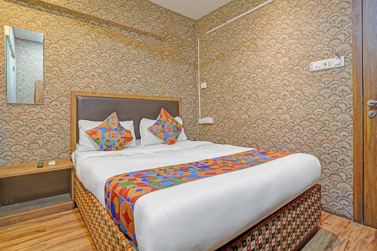A neatly made bed with a colorful geometric-patterned bedspread and pillows, a wooden headboard, a nightstand with a lamp, and a patterned wallpaper.