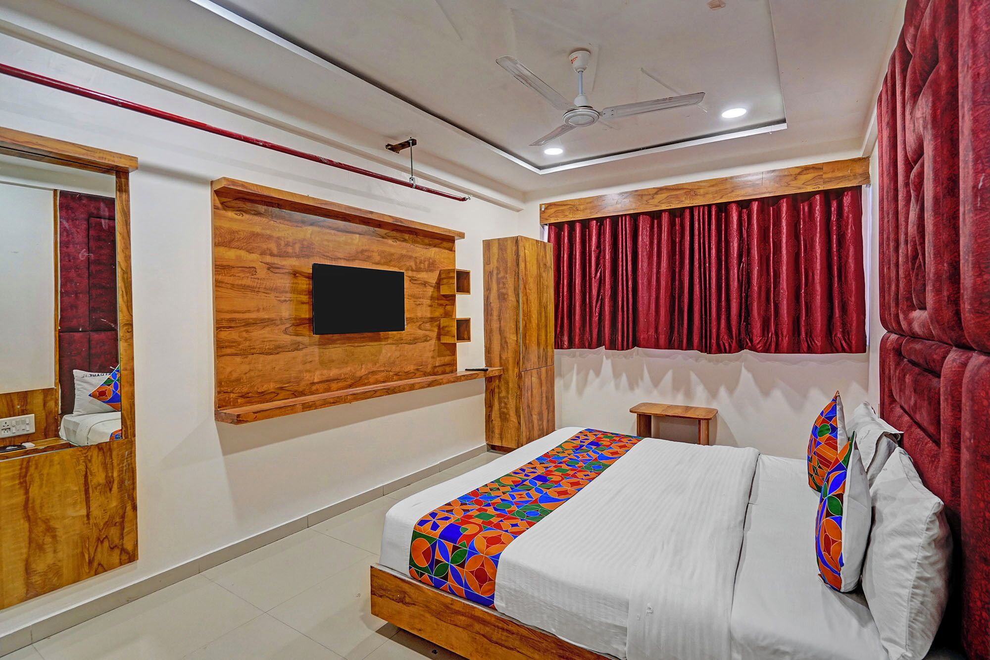 Un dormitorio con cabecera de madera, cama con colcha geométrica colorida, televisor montado, ventilador de techo y cortinas rojas.